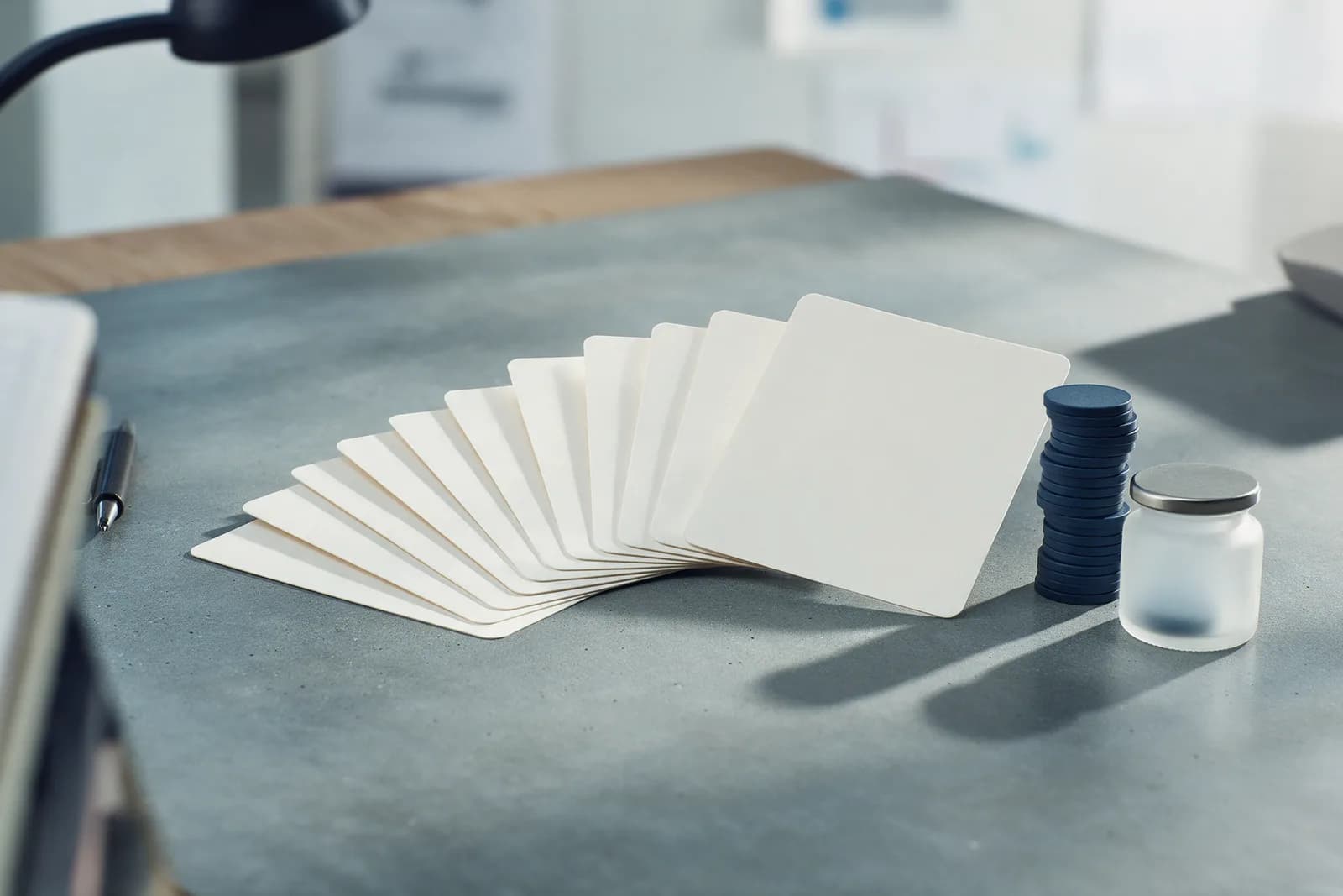 A fan of blank budget cards growing from small to large on a grey desk, ending at a stack of navy tokens and a frosted jar.