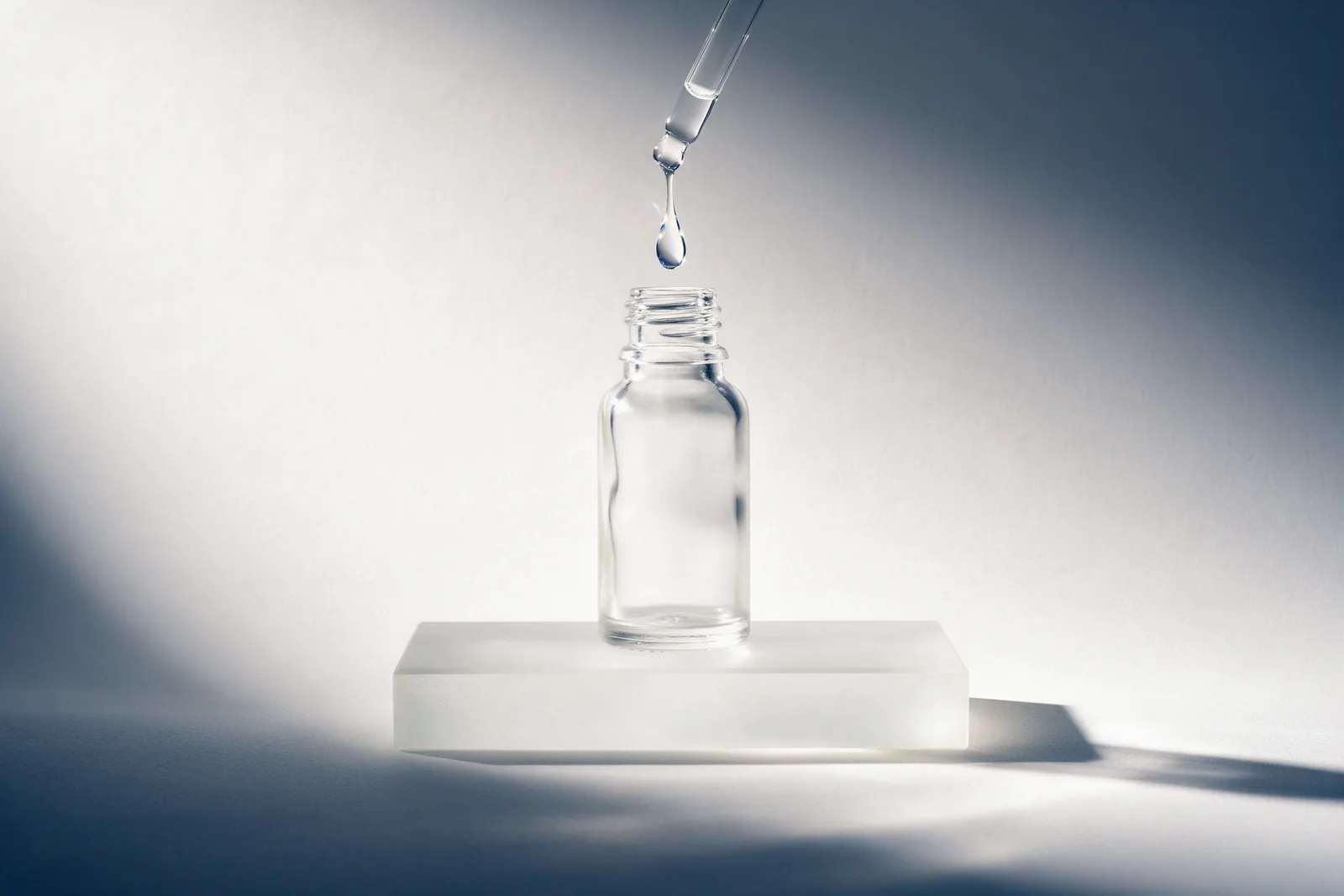 A clear drop falls from a dropper into a frosted glass bottle on a frosted riser, deep blue shadow across a pale backdrop.