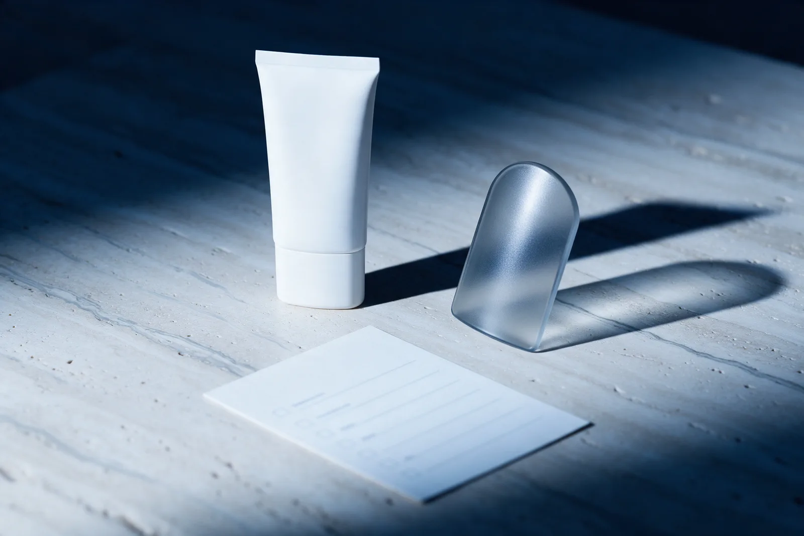 An unlabelled sunscreen tube beside a frosted glass shield and a blank checklist card on pale stone in hard cool light.