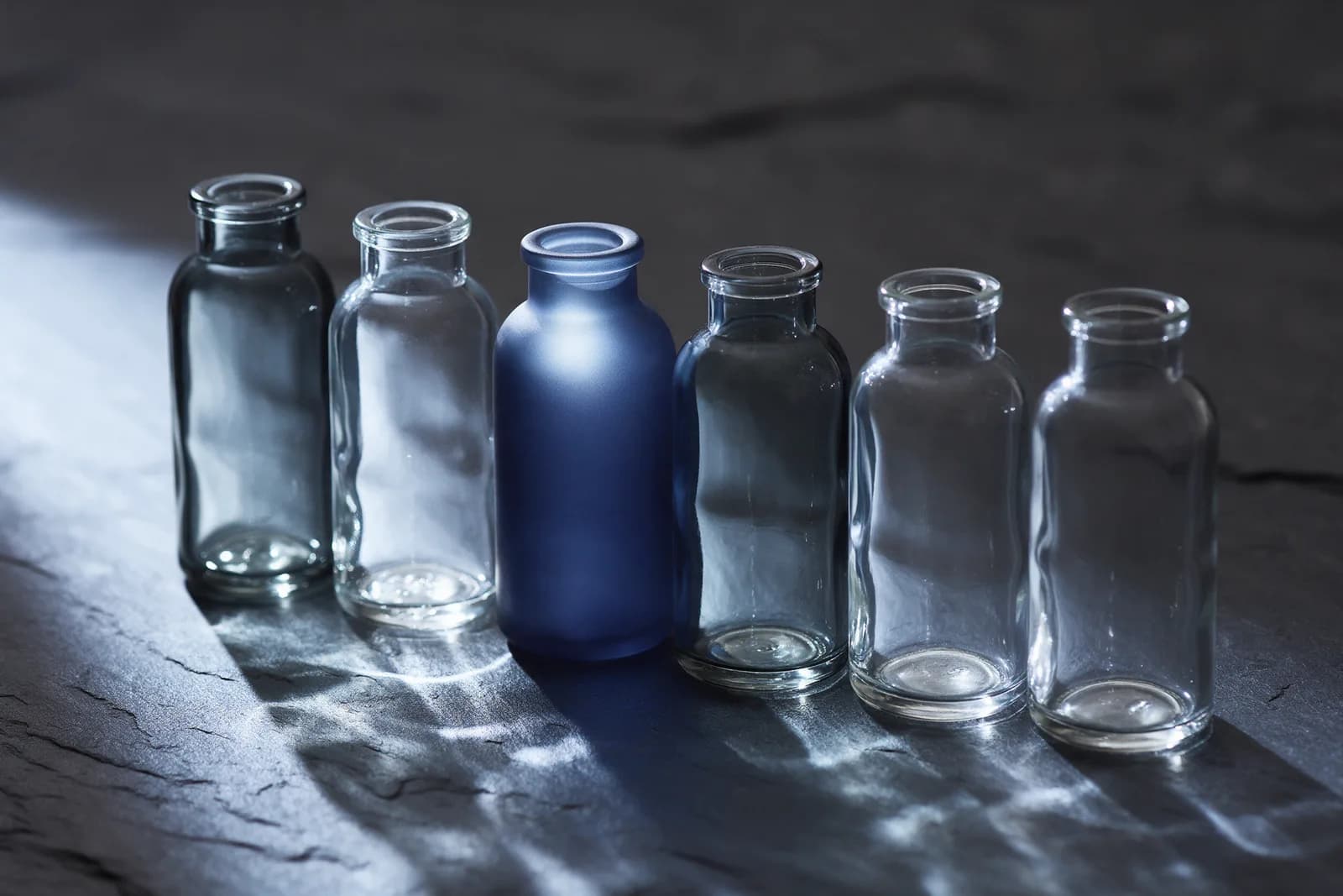 Six glass bottles in a row on slate, five clear and one in frosted navy glass, in cool north light.