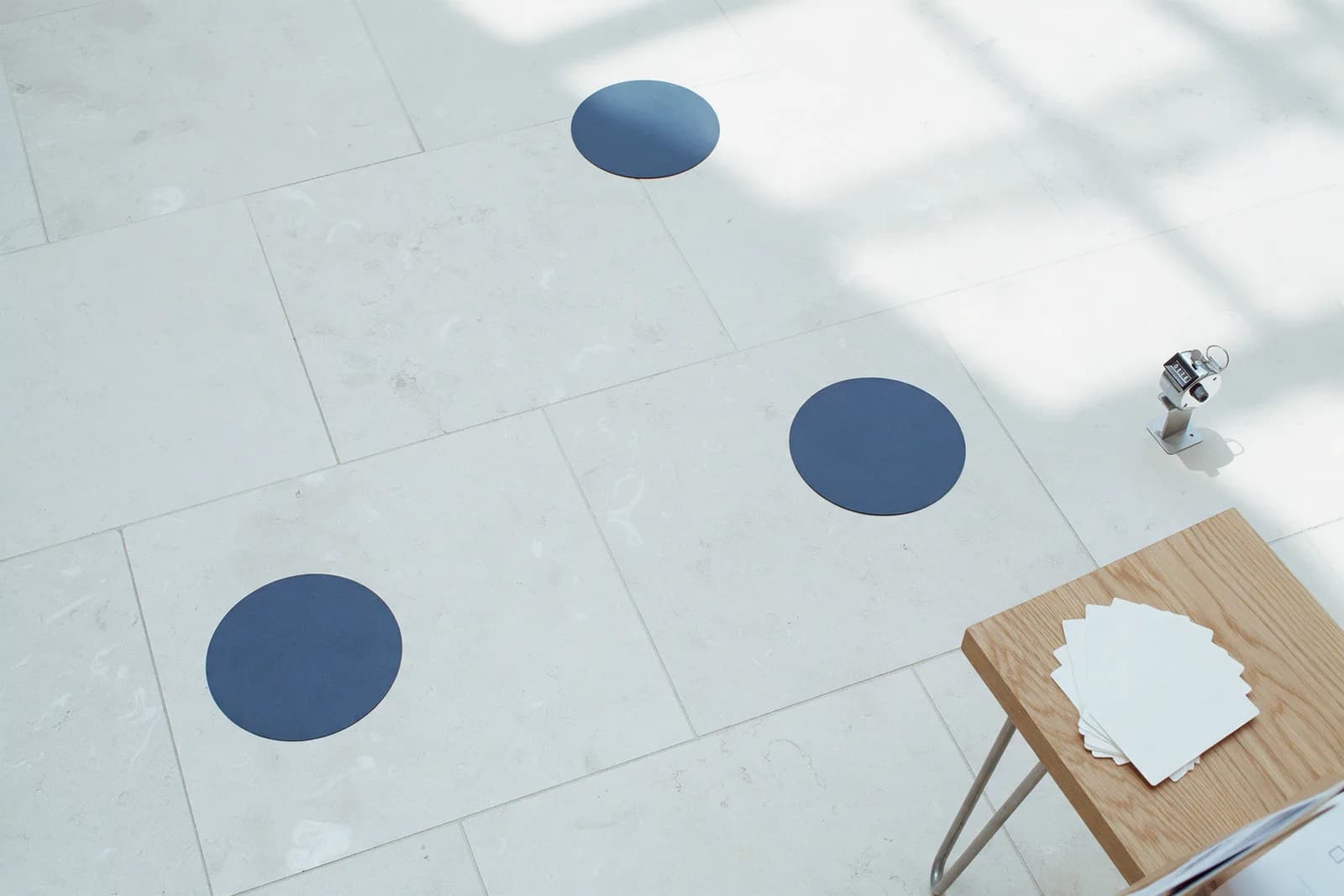 An empty pale stone floor seen from above with three navy floor markers, a tally counter and blank cards on a side table.