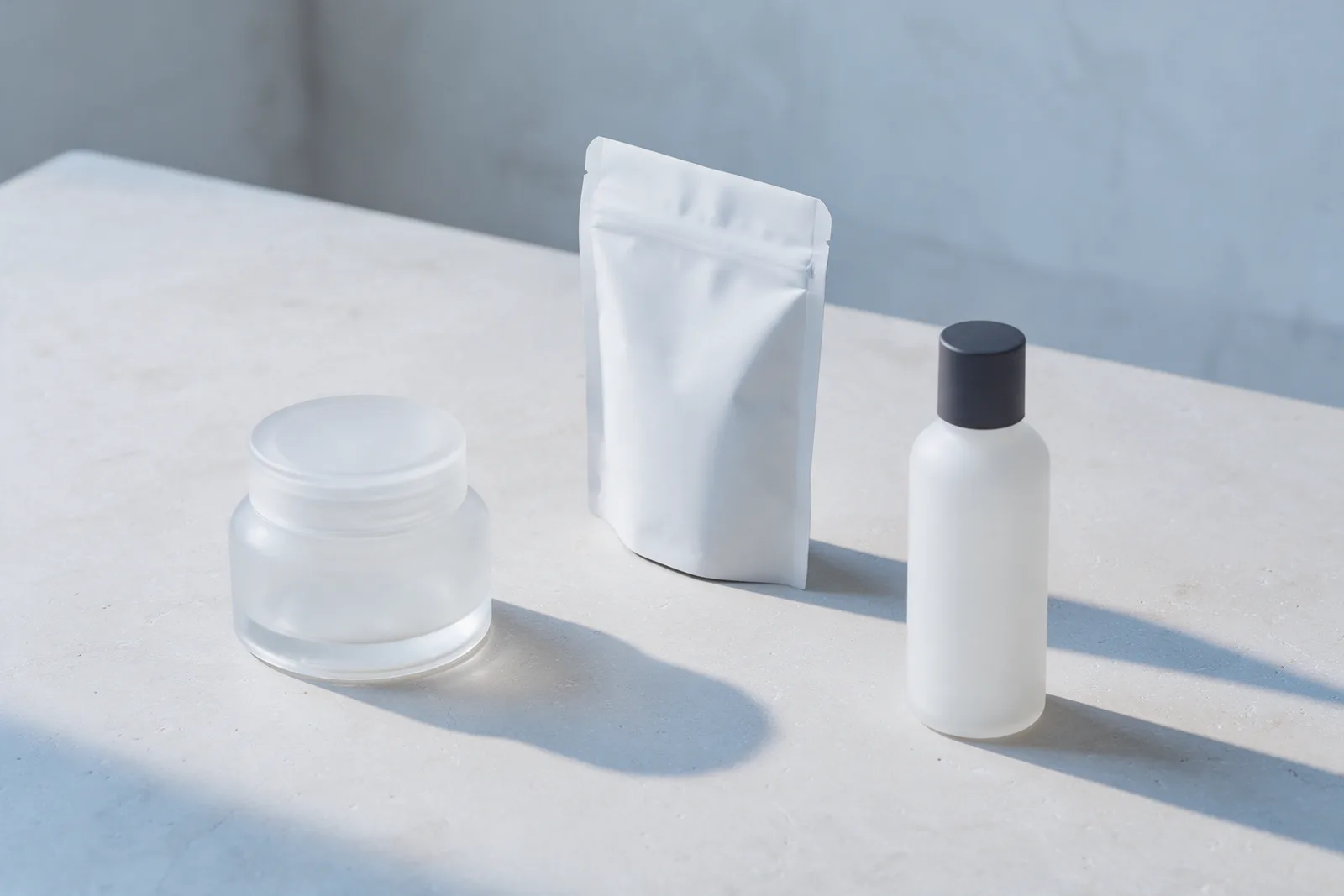 A glass jar, a white pouch and a white bottle with a dark cap lined up on pale stone under the same cool light.
