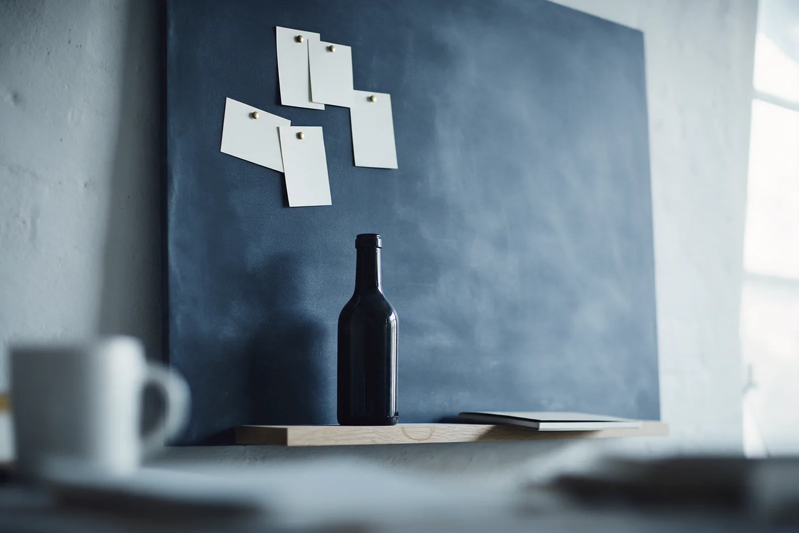 A small cluster of blank cards pinned to a navy board above a dark unlabelled bottle on a narrow shelf, in cool light.