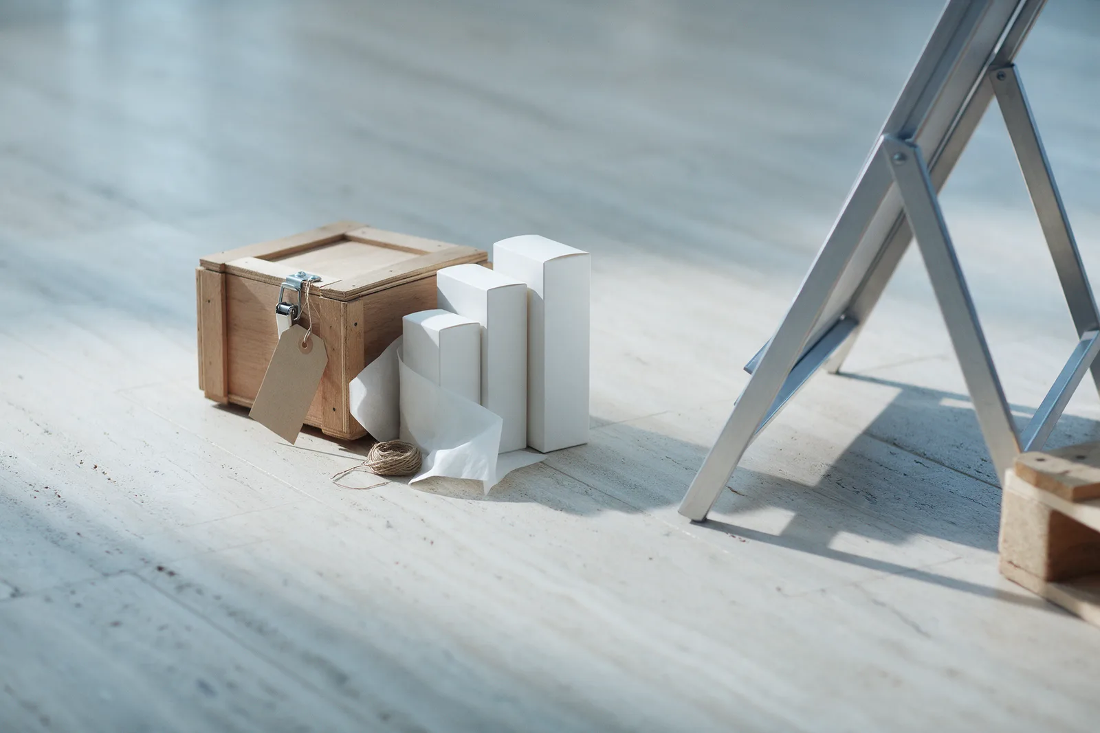 Unlabelled white skincare boxes beside a small crate with a blank tag and a folding display stand on a pale stone floor.