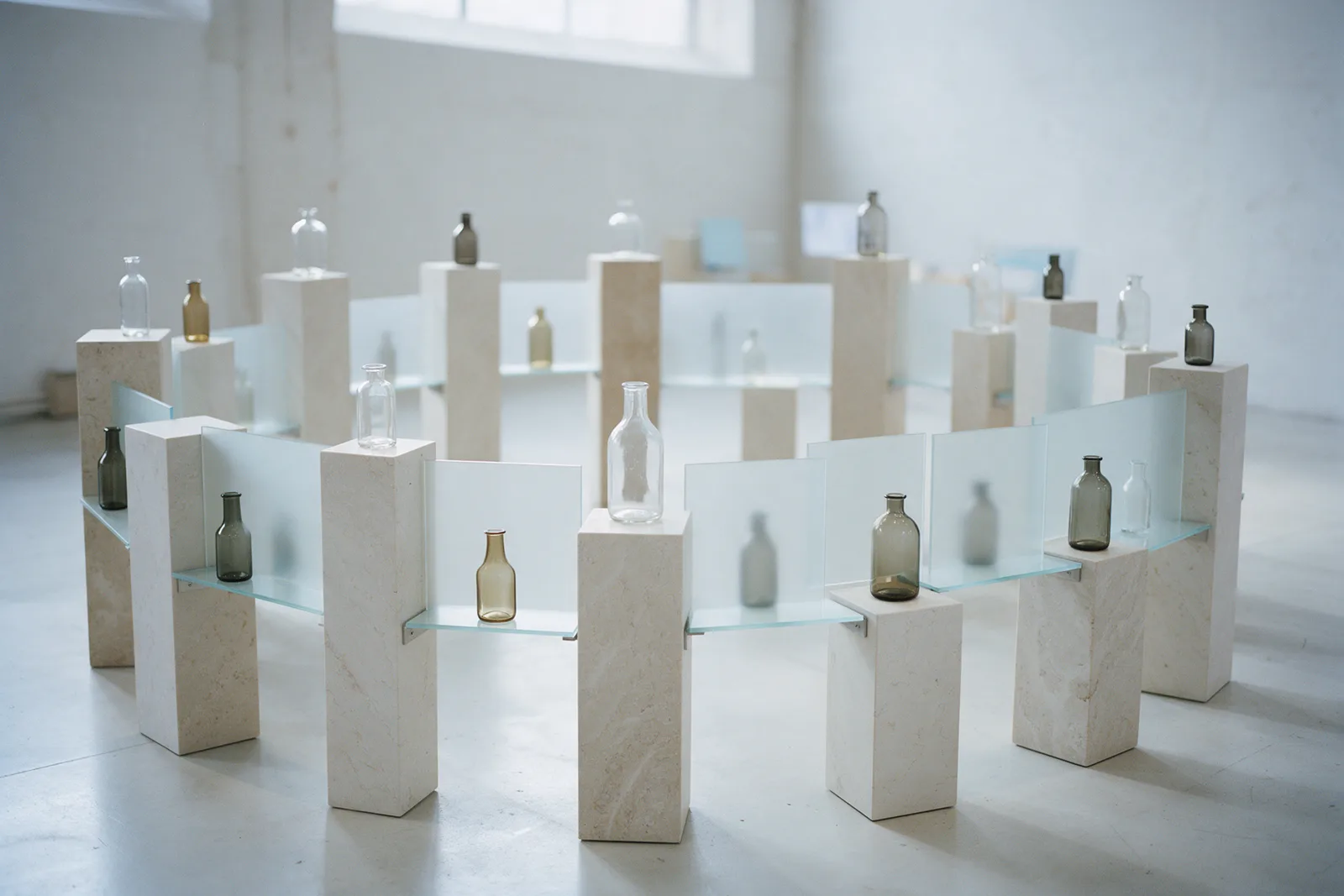 A curved ring of pale stone plinths linked by frosted glass shelves, each with a small unlabelled bottle, in a white gallery.