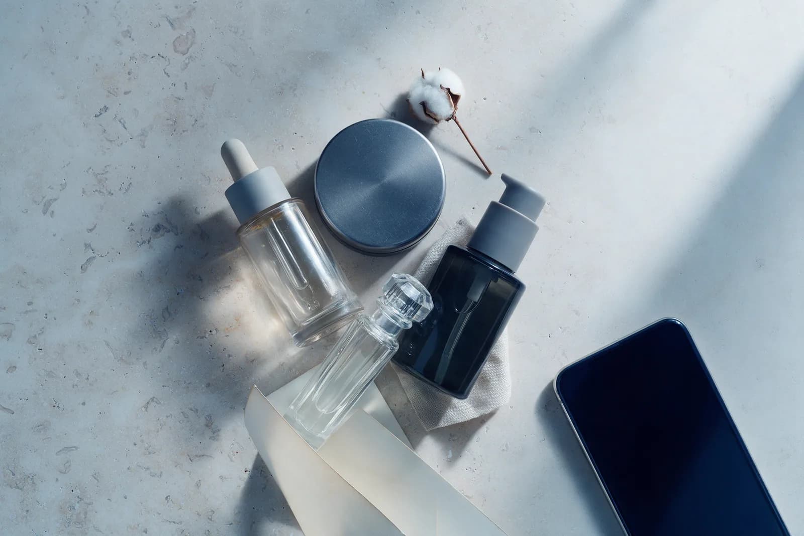 A flat lay of a dropper bottle, a steel compact, a pump bottle and a fragrance vial on pale stone beside a dark blank phone.