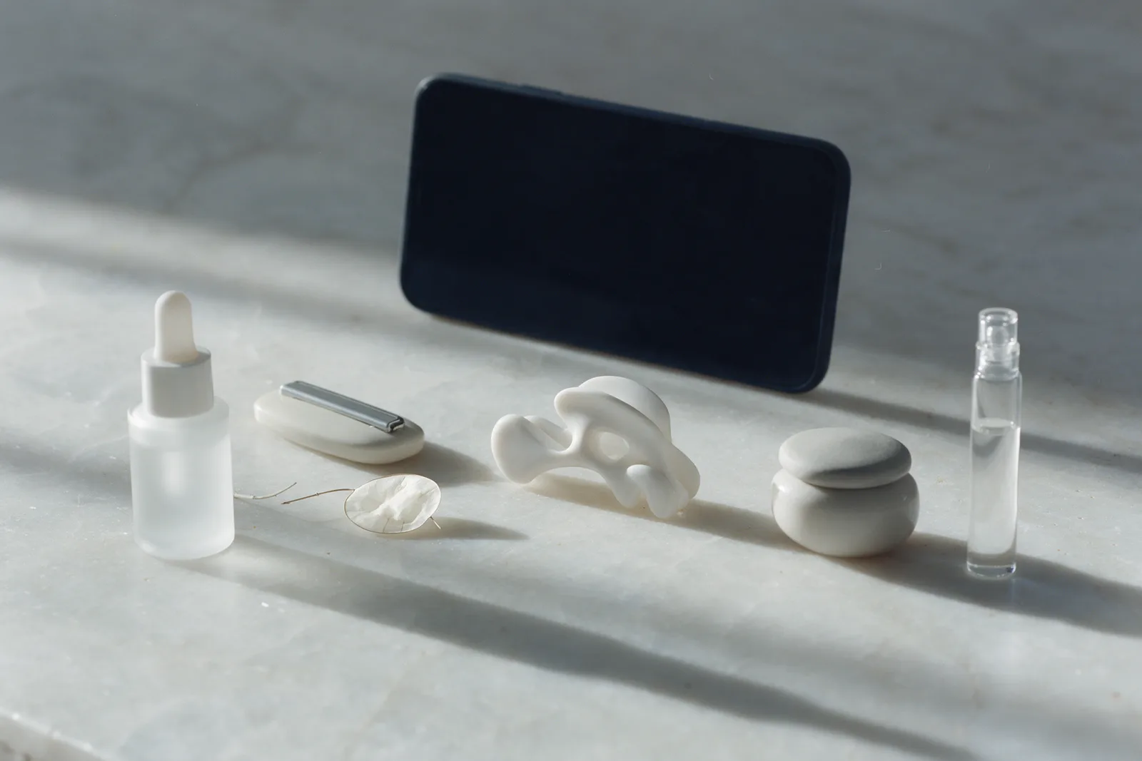 A dropper bottle, a compact, a hair clip, a small jar and a fragrance vial in an arc on pale stone before a dark blank phone.