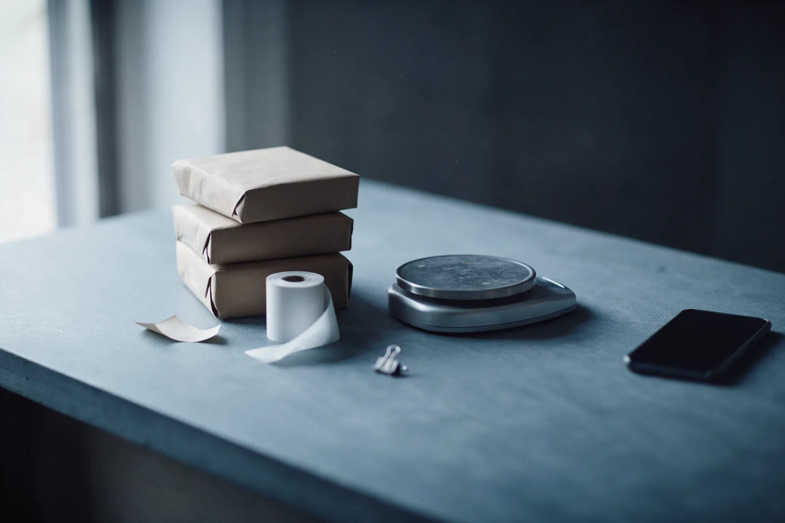 A stack of plain paper-wrapped parcels beside a blank receipt roll, a small steel scale and a dark phone on slate.