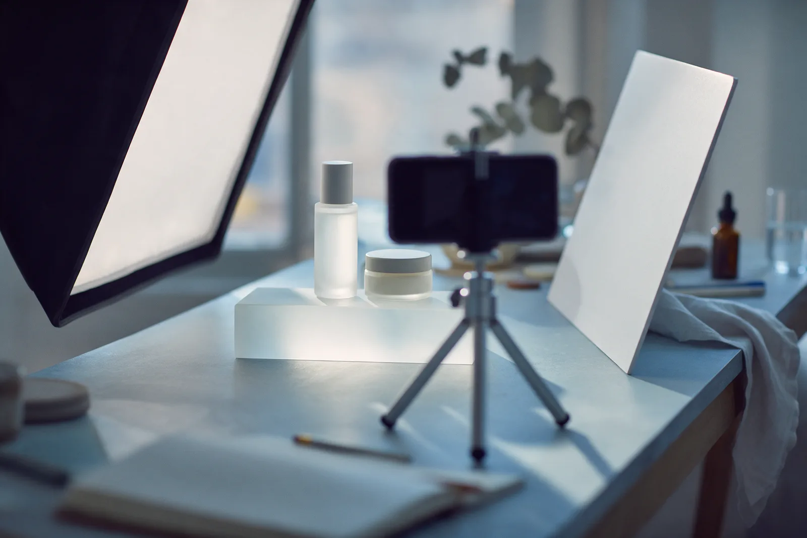 A phone on a small tripod aimed at a bottle and jar on a frosted glass riser, a softbox and white reflector around it.