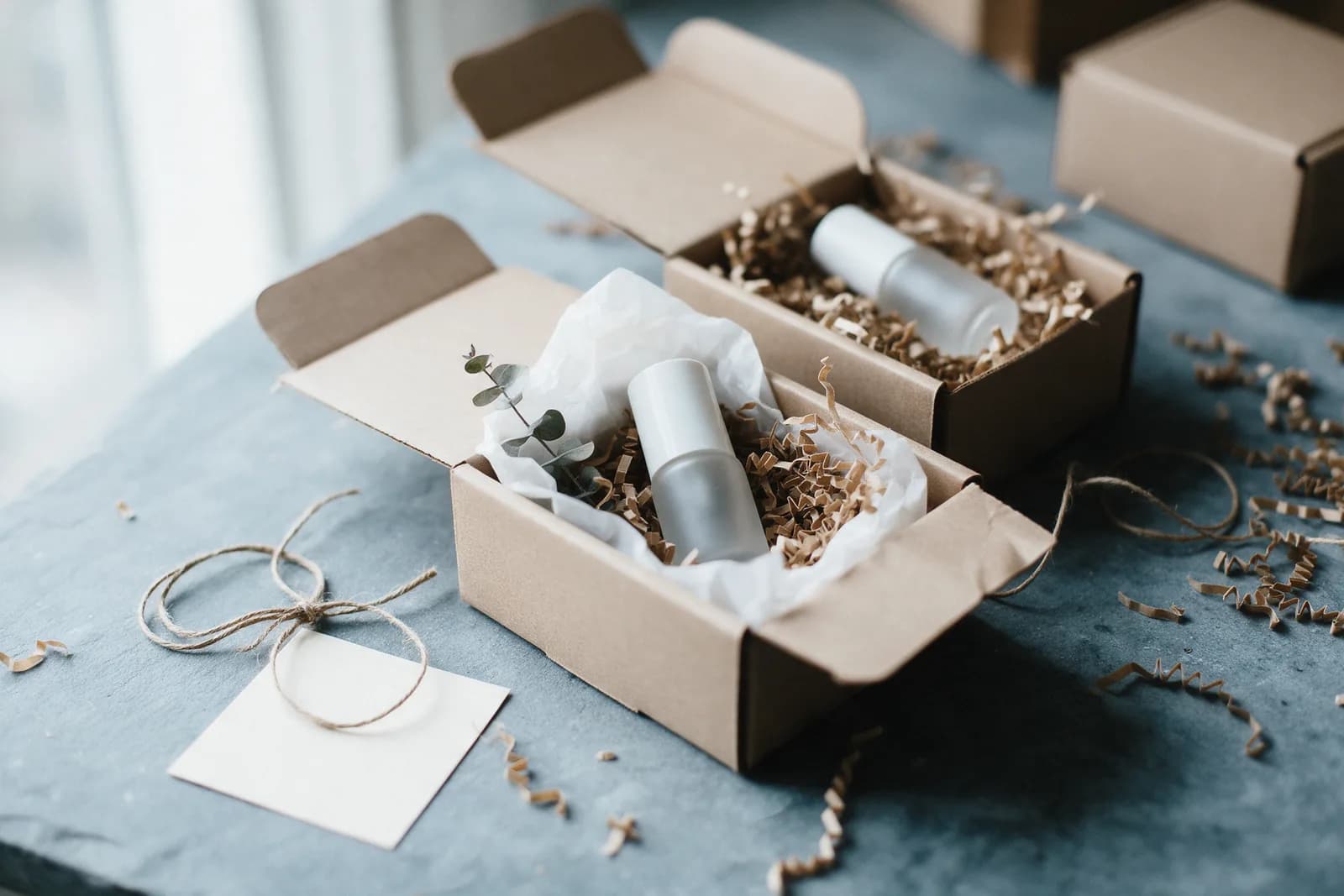Two open kraft boxes on a slate table holding frosted skincare bottles nested in tissue and shred, with twine and a blank card.